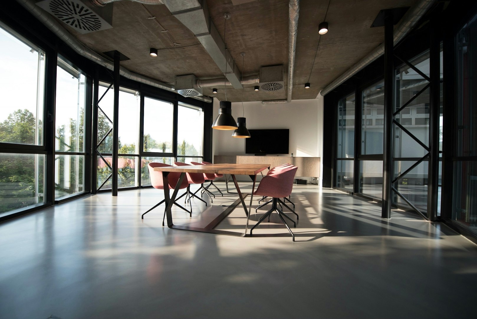 A modern kitchen featuring sleek black and gray cabinetry, a large white countertop, and an island with a wooden table and chairs. The space is illuminated by recessed lighting and has large windows letting in natural light, with decorative plants and dishes on display.